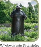 A bronze Old Woman with Birds statue standing among garden plantings