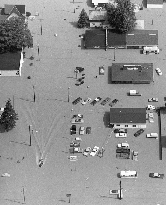 A black-and-white aerial photograph of a flooded town area with cars and buildings